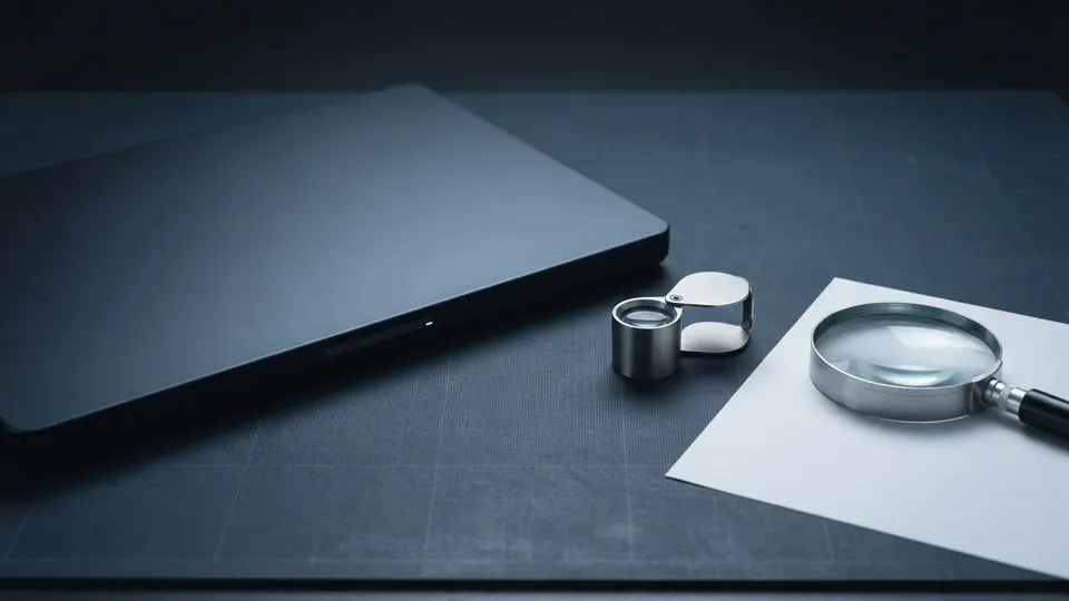 Closed laptop beside a steel jeweller's loupe and a magnifying glass lying on a blank sheet of paper