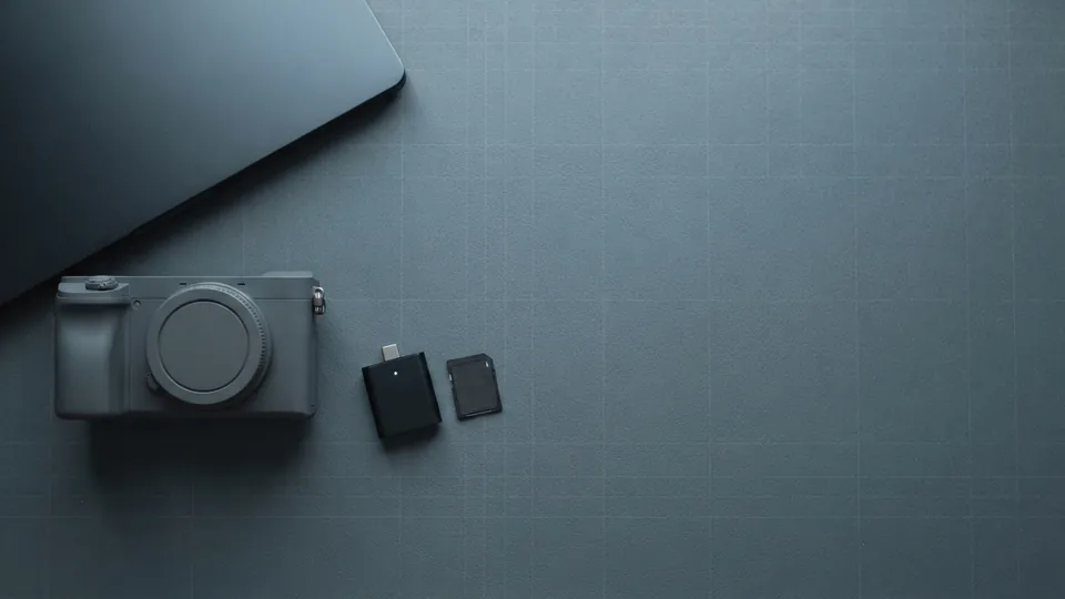 Compact camera, a memory card and a small card reader laid out beside the corner of a closed laptop on a slate mat