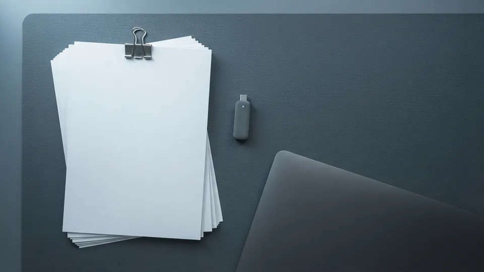 Stack of blank paper held with a binder clip beside a small USB drive and a closed laptop