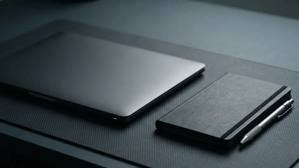 Slim closed laptop next to a dark notebook and a steel pen on a gridded bench mat