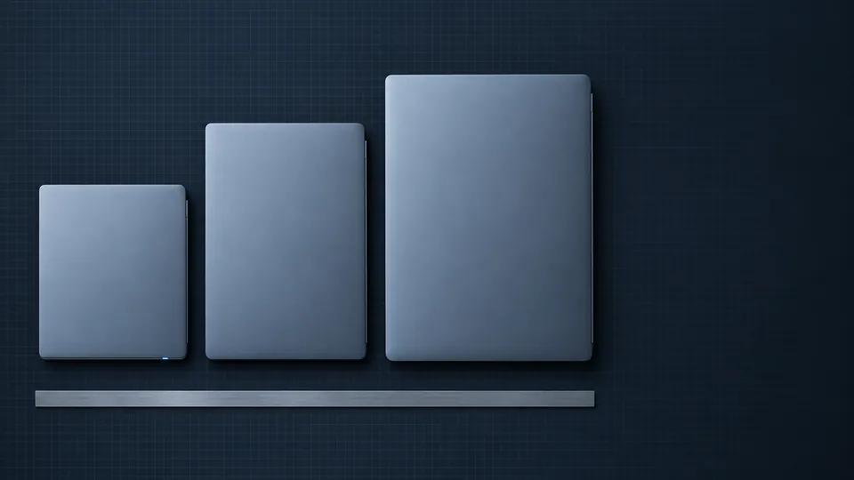 Three closed laptops of different sizes laid side by side above a steel ruler on a slate test bench, seen from overhead